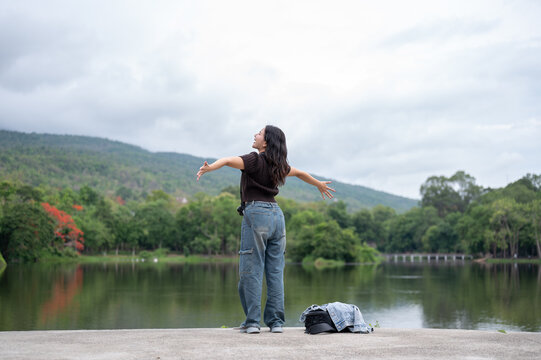 Back view shot of asian woman opening both arms while standing beside the lake and mountain in park.