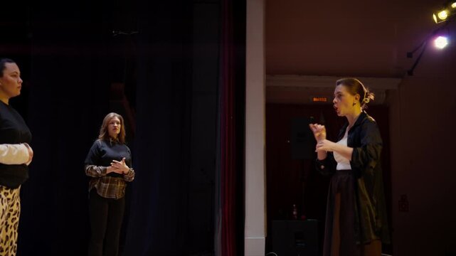 Theater rehearsal on dimly lit stage with director instructing two performers during acting practice, emphasizing expression, movement, and collaborative performance