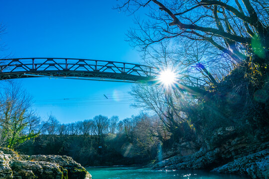 A metal bridge spans the turquoise waters of Martvili Canyon, with bare trees, rocky canyon walls, and bright sunlight creating a dramatic winter scene in western Georgia.