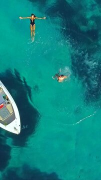 Vertical aerial of a man performing a backflip jump from a boat into the clear turquoise sea, woman relaxing in the water, carefree summer fun couple lifestyle