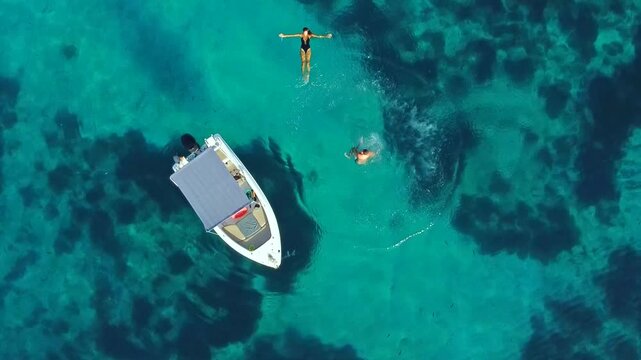 Aerial of a man performing a backflip jump from a boat into the clear turquoise sea, woman relaxing in the water, carefree summer fun couple lifestyle