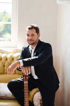 Handsome man in suit playing guitar while sitting on sofa at home