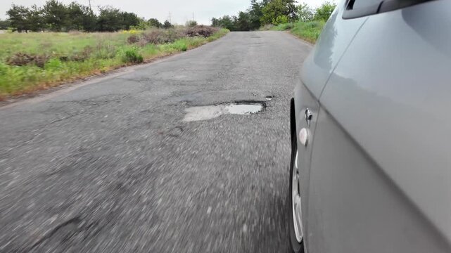 Slow-motion video shot from a moving silver car's perspective on a bumpy asphalt road in the countryside. The car's body 