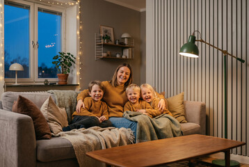 Obraz premium A joyful multi-generational family scene: a mother and three young children pose happily together on a plush living room sofa at home during the evening.