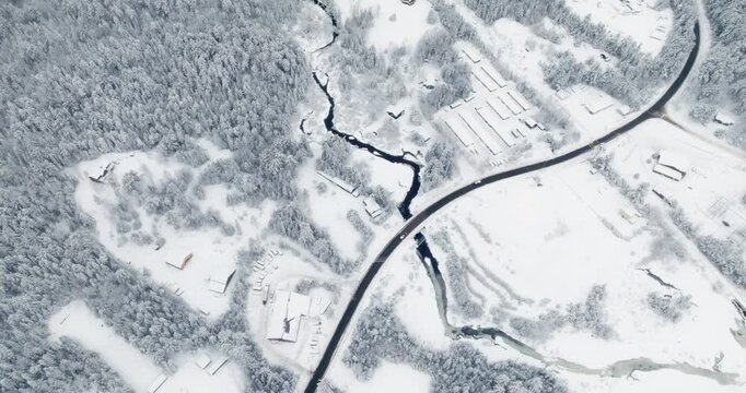 Drone flies forward over Lake Placid during fresh snowfall, revealing a winding river cutting through snow-covered forest, intersected by a dark road and scattered rural buildings.