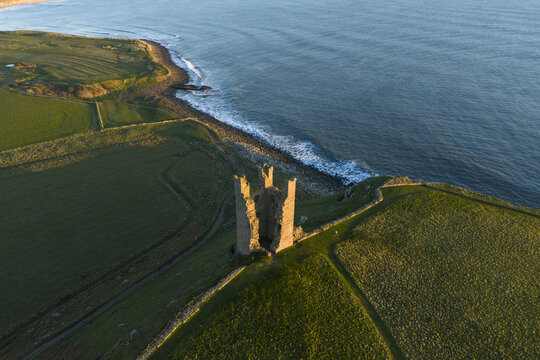 Aerial view of the ruined Dunstanburgh Castle on a grassy headland overlooking the blue sea during golden hour Craster, England, United Kingdom.
