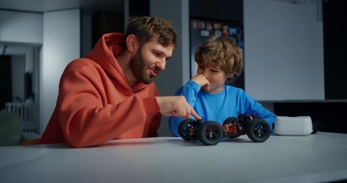 Father and young son smiling while assembling and testing a remote control toy car together at a kitchen table during a cozy evening at home