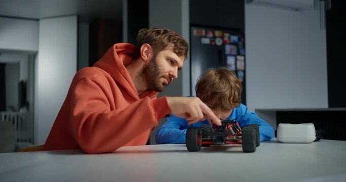 Father and young son collaborating at a kitchen table, examining and adjusting a small remote controlled toy car during a focused learning activity
