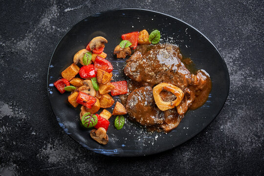 Traditional Italian Osso Buco served with roasted potatoes, mushrooms, and bell peppers on a black plate. Flat lay of a gourmet braised veal shank meal on a dark stone background.