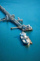 Fototapeta premium A cargo ship is docked at an industrial port. The blue water surrounds the pier with many structures used for loading and unloading
