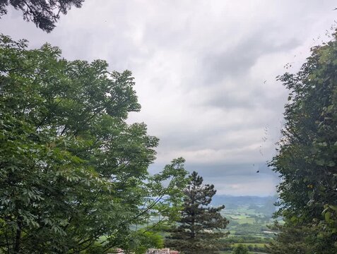 Veduta naturale incorniciata da alberi, con apertura sulla valle e colline verdi in lontananza sotto un cielo nuvoloso. Atmosfera tranquilla e immersiva, tipica dei paesaggi italiani.