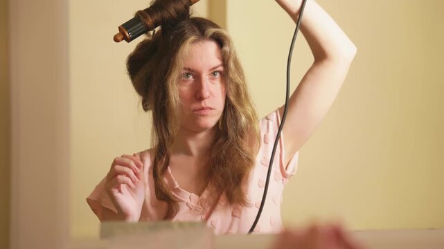 Focused young woman using a modern hair styling tool to create voluminous curls, looking at her reflection in the mirror during her daily beauty routine in a bathroom with soft lighting