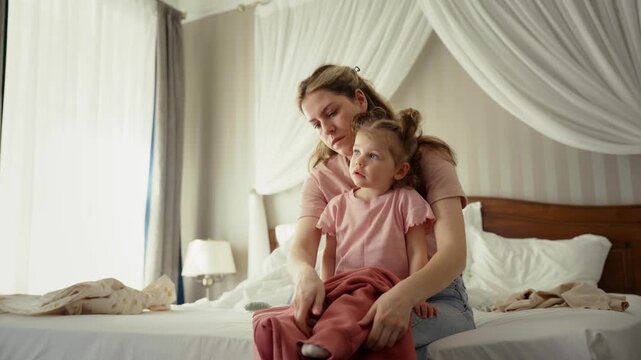 Loving mother sitting on a bed and carefully putting pants on her little daughter while getting ready for the day, showcasing a quiet moment of family care and morning routine at home