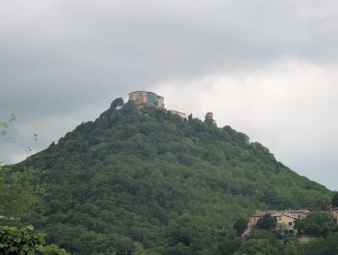 Collina ricoperta di vegetazione, sormontata da un&rsquo;imponente struttura storica in pietra. Atmosfera suggestiva e panoramica, con piccoli edifici immersi nel verde ai piedi del promontorio