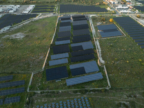 Aerial view of a large solar farm with rows of photovoltaic panels on green fields near an industrial zone in Augusta, Sicily, Italy.