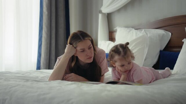 Young loving mother telling a story from a picture book to her adorable little daughter while relaxing together on a cozy bed, enjoying a quiet moment of bonding and early childhood education