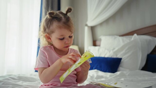 Toddler girl with cute hairstyle sits on a white bed at home, focused on peeling colorful stickers from a book, developing fine motor skills during quiet playtime
