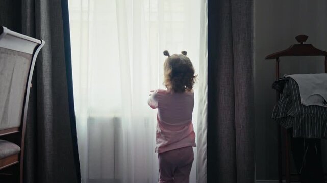 Back view of a small child with curly hair running towards a large window and opening the white curtains to look outside, standing in a cozy room at home with bright daylight shining through