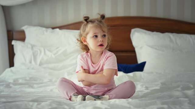Cute toddler with a funny hairstyle sitting on a large bed in her pajamas, crossing her arms with a stubborn expression before changing her mind and looking away with curiosity