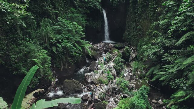 Drone footage flying over a narrow tropical gorge with a waterfall surrounded by dense vegetation and visible plastic waste in Bali, Indonesia.