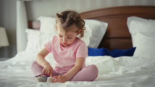 Cute little girl with curly hair sitting on a big bed in the morning and playing with a modern mobile phone. Child watching cartoons or playing educational games on a digital device at home