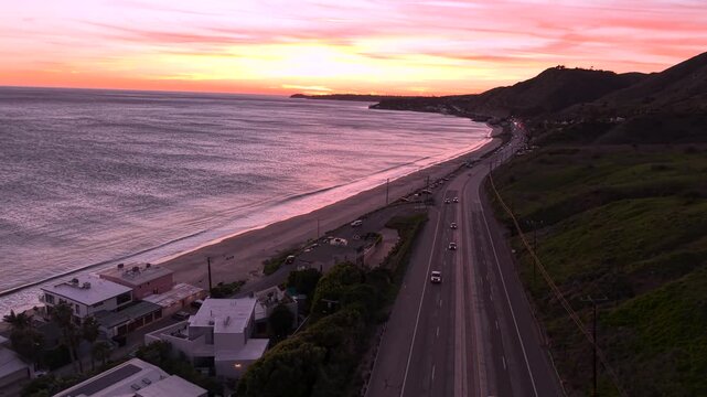 EPIC view driving to Los Angeles, on Malibu coast along PCH Highway 1, with a beautiful pink sunset reflecting on Pacific Ocean water, waves crashing in from the glowing California horizon.