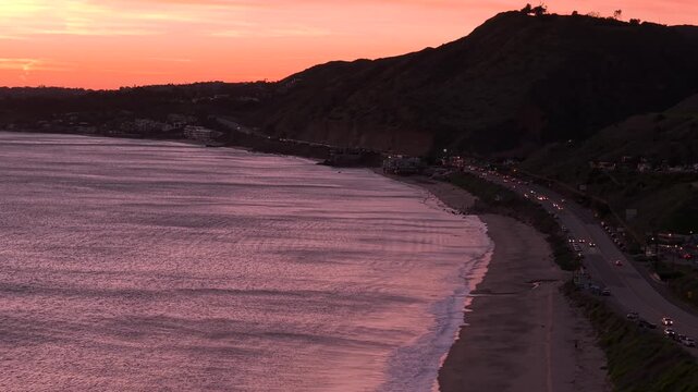 California's West Coast beach shoreline at golden hour, warm light and pink skies spreading across sand and surf in a dreamy Pacific Ocean landscape. Cars drive along PCH Highway with headlights on.