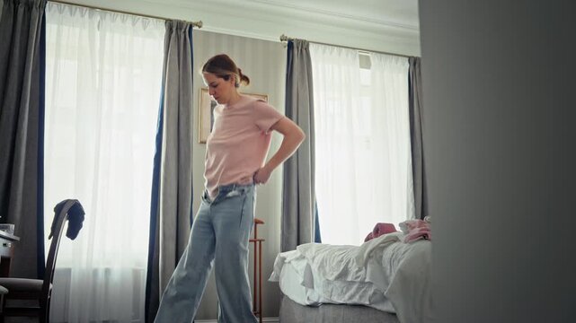 Young woman getting dressed in her bright bedroom in the morning, struggling to put on a pair of tight blue jeans. Daily routine and lifestyle concept as she prepares for the day