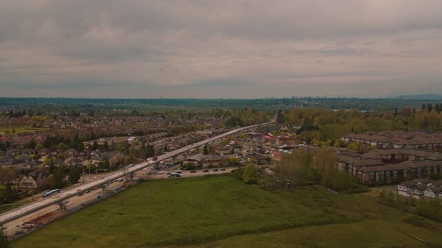 Suburban highway traffic flowing alongside elevated concrete guideway construction site surrounded by sprawling residential housing developments under dark overcast clouds right orbit