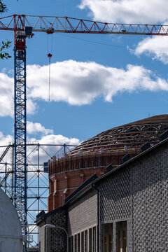 A crane rises over a construction site as sky and clouds open above an industrial roof, scaffold and patterned wall around the structure