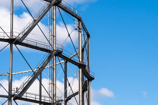 A steel gasholder framework opens against blue sky, where industrial beams and heritage structure reveal landmark geometry above the district