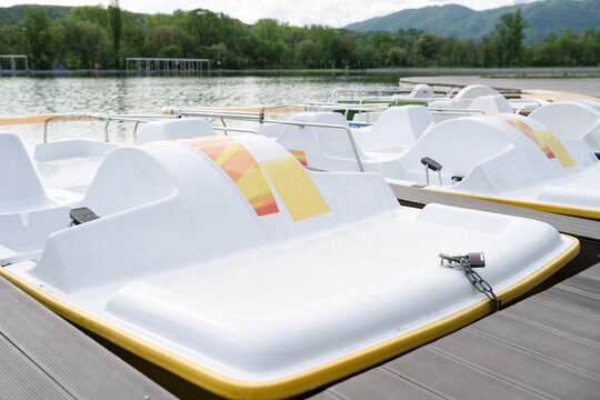 A row of modern white paddle boats parked at a wooden pier on a calm lake with green forest and mountains in the background.