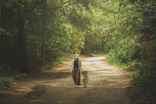 Happy Asian mother and her young daughter walking hand-in-hand on a dirt road through a lush green forest. Serene family bonding and nature exploration during a summer hiking trip. Authentic lifestyle