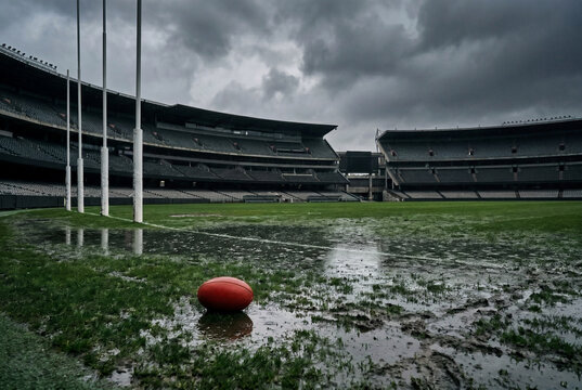 Australian rules football ground with a red football lying in puddles and mud under a dark, cloudy sky