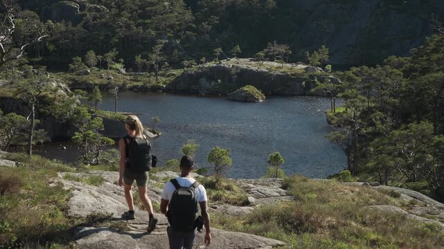 Hikers walk along a small mountain lake in Norway, pause at the shore to enjoy the scenery, then continue their hike through a peaceful alpine landscape.