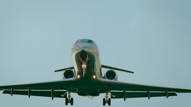 Private jet flying with landing gear extended under clear pale sky. Executive aircraft approaching runway with twin rear engines and lights glowing. Business airplane descending in daylight with