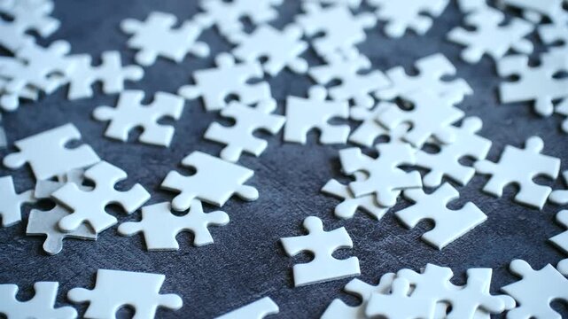 Numerous blank white jigsaw puzzle pieces lying scattered on dark gray table, creating conceptual scene of problems, solutions, connection, teamwork challenges. White jigsaw puzzle pieces on table