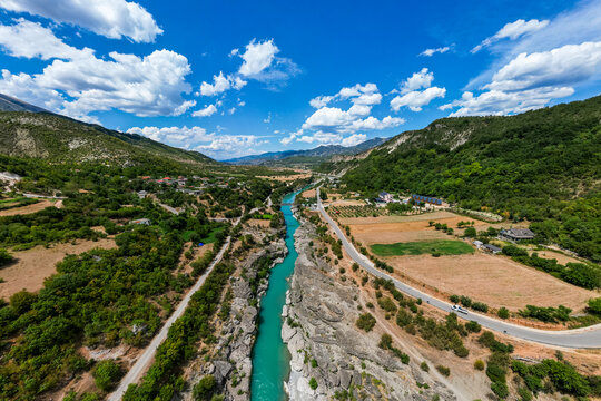 Aerial view of the stunning turquoise Vjosa River near Permet, Albania, featuring a narrow bridge spanning a rocky gorge with traditional mountain villages and rolling hills in the background.