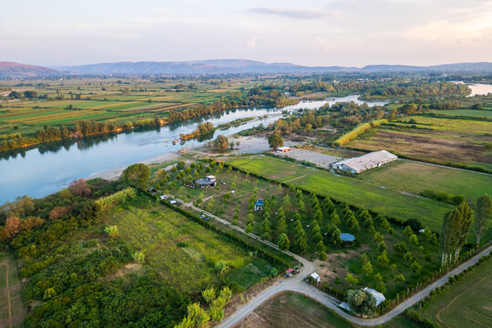 Aerial panorama of a small, green eco-campsite nestled along the banks of the Buna River in Northern Albania, featuring lush agricultural fields and mountain views under a soft sunset sky.