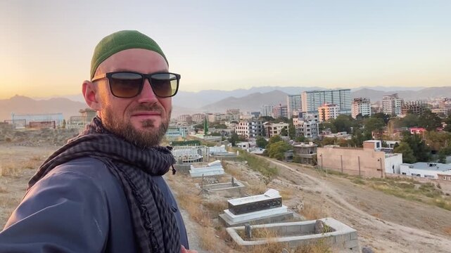 western tourist traveler visit kabul taking selfie at city viewpoint. man with sunglasses & shemagh scarf overlooking urban afghanistan landscape. travel vlogger, tourism, & exploration