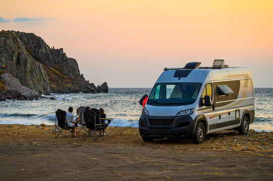 Family relax in folding chairs next to their modern campervan on a rugged beach in Limnos, Greece, enjoying a sunset view of the Aegean Sea during an off-grid motorhome road trip.