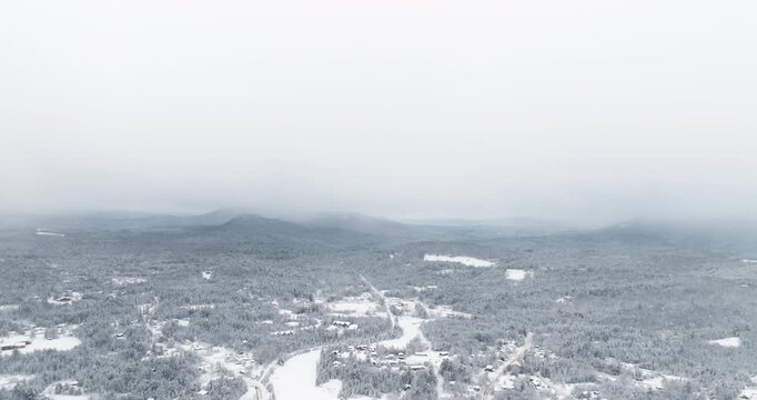 Drone fly in across the snow-covered Lake Placid valley in New York as low winter clouds drift through the Adirondack Mountains and fresh snowfall blankets forests and homes below.