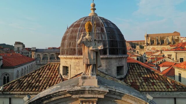 Drone aerial close-up of Saint Blaise Church baroque dome with bishop statue in golden morning light overlooking Dubrovnik old town red rooftops