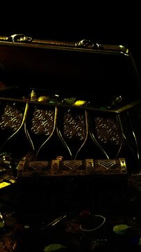 A treasure chest is open, revealing various gems and gold coins inside. The setting is dark, highlighting the shiny objects. The chest has intricate designs and is made of dark metal.