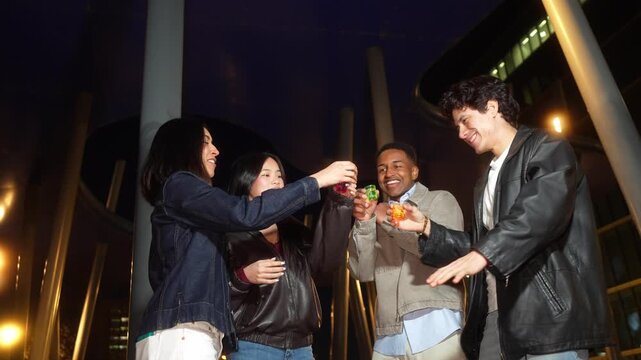Slow motion of diverse young friends toasting, drinking shots, and dancing together with city lights in the background at night