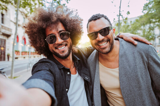Two smiling latin american men wearing sunglasses taking a selfie together on an urban street during sunset.