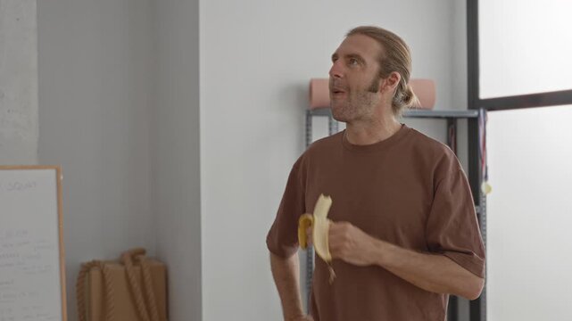Man holding a peeled banana in a yoga studio, wearing a brown t shirt and hair bun, chewing while looking upward beside mats and trophies; post workout contemplation.