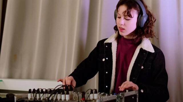Young girl in casual jacket works carefully at a mixing console with headphones on, creating modern electronic music and developing creative technical skills.