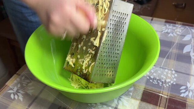 Grating raw potatoes for potato pancakes or hash browns; the grated potatoes fall into a green bowl, close-up