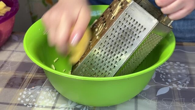 The process of grating raw potatoes on a metal grater into a green bowl, preparing ingredients for traditional dishes